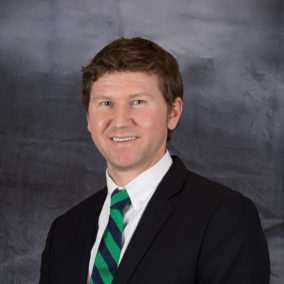 A photograph of Keven DuComb, wearing a suit, standing in front of a neutral background.